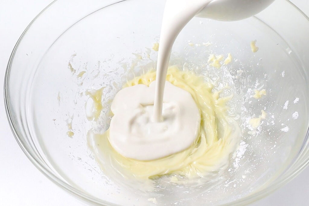 A thick, creamy liquid is being poured into a clear mixing bowl with the cream cheese mixture.