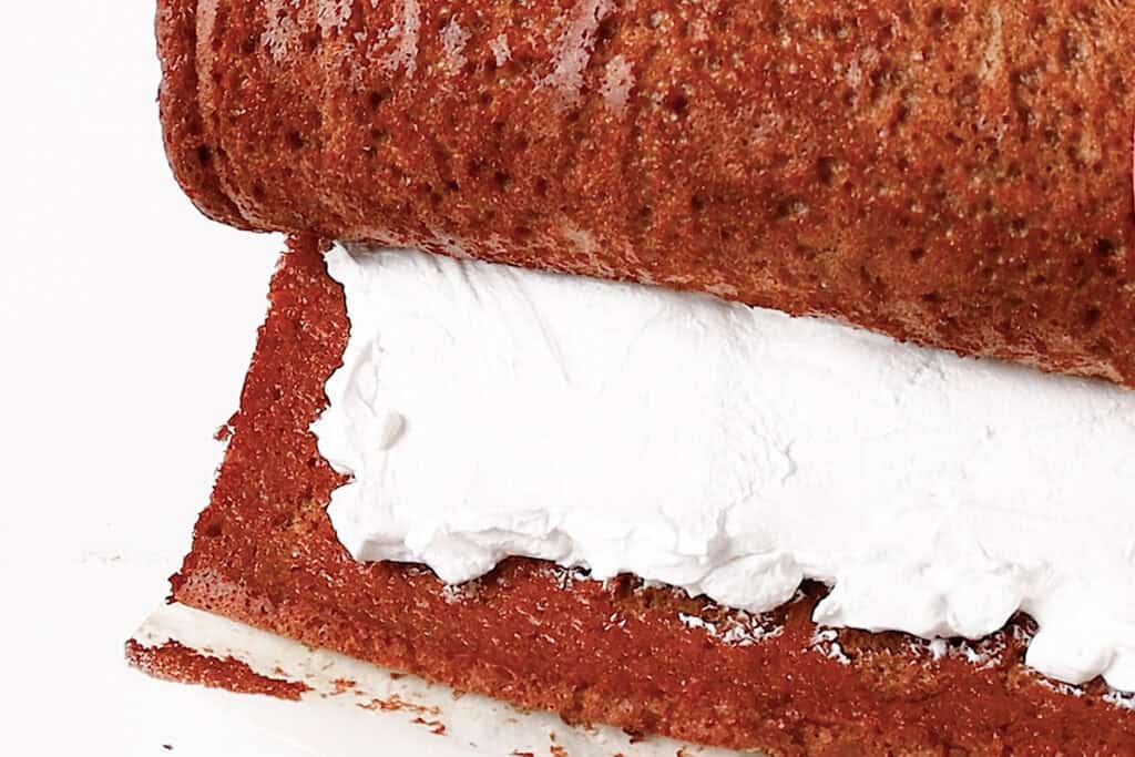 A close-up of a Pumpkin Swiss Roll being prepared. The image shows a brown, textured cake partially unrolled, revealing a thick layer of white cream filling inside. The cake is on a white surface.