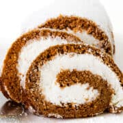 Close-up of two slices of a Pumpkin Swiss Roll with a rich brown sponge and a white cream filling, dusted with powdered sugar. They are placed on a white plate, next to an ornate serving spatula. The background is white.