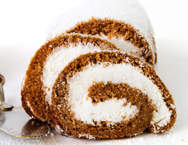 Close-up of two slices of a Pumpkin Swiss Roll with a rich brown sponge and a white cream filling, dusted with powdered sugar. They are placed on a white plate, next to an ornate serving spatula. The background is white.