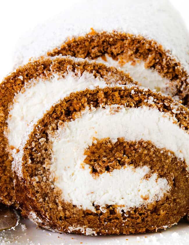 Close-up of two slices of a Pumpkin Swiss Roll with a rich brown sponge and a white cream filling, dusted with powdered sugar. They are placed on a white plate, next to an ornate serving spatula. The background is white.