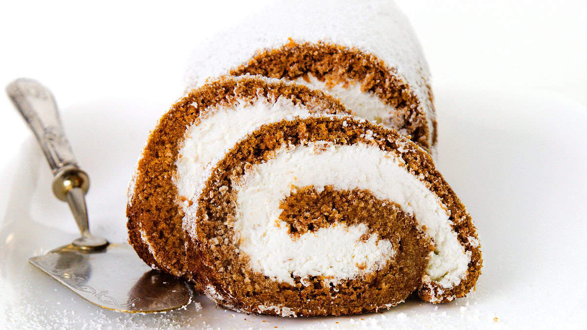 Close-up of two slices of a Pumpkin Swiss Roll with a rich brown sponge and a white cream filling, dusted with powdered sugar. They are placed on a white plate, next to an ornate serving spatula. The background is white.