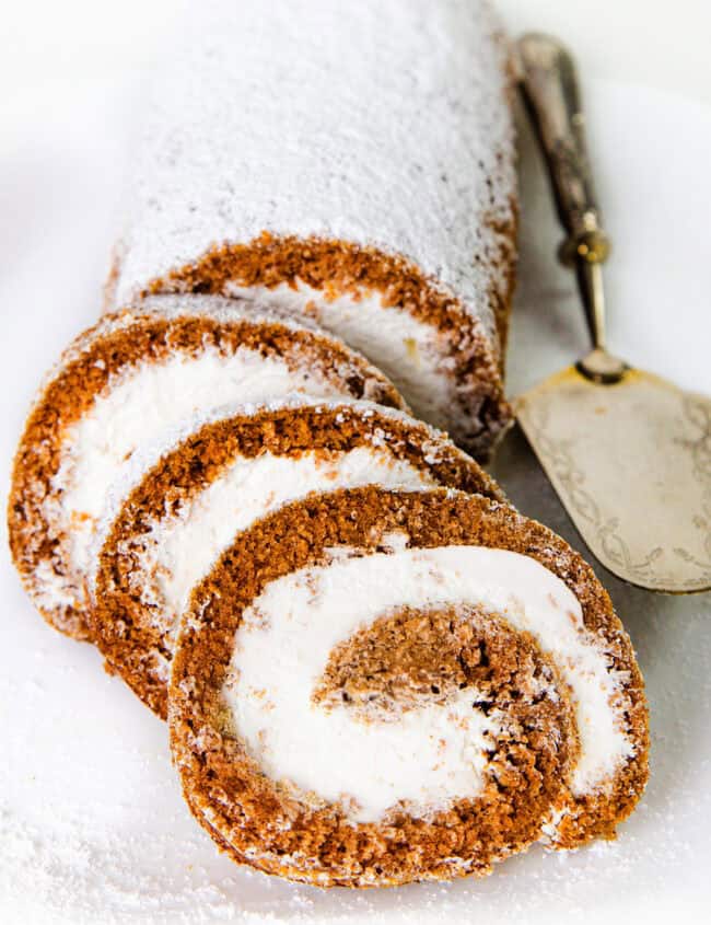 A sliced Pumpkin Swiss Roll cake is displayed on a white plate. The cake appears light brown with a generous dusting of powdered sugar and has a white, creamy filling swirled through each slice. A vintage serving spatula lies beside the cake.