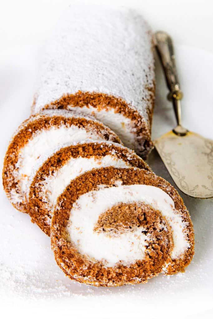 A sliced Pumpkin Swiss Roll cake is displayed on a white plate. The cake appears light brown with a generous dusting of powdered sugar and has a white, creamy filling swirled through each slice. A vintage serving spatula lies beside the cake.