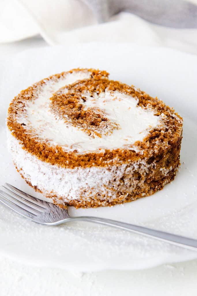 A close-up shot of a single slice of Pumpkin Swiss Roll with a creamy white filling, lightly dusted with powdered sugar. The slice is served on a white plate with a metallic fork resting beside it. The background features a white and gray cloth.