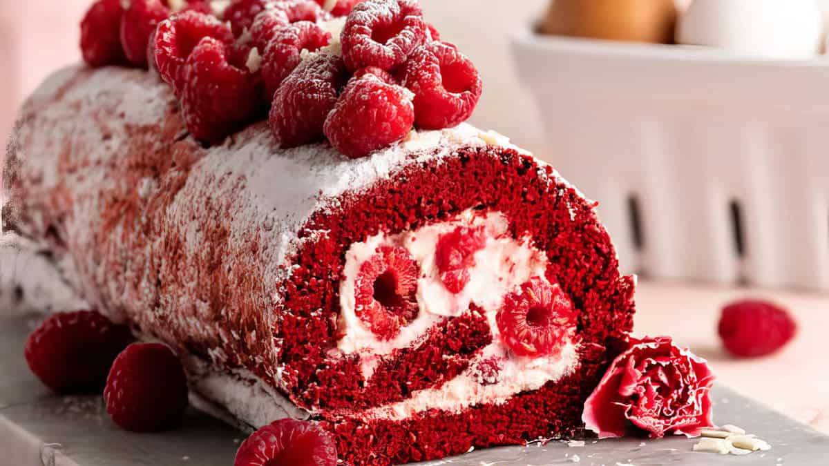 A delicious red velvet cake roll, filled with creamy white frosting and fresh raspberries, is placed on a marble counter. The roll is garnished with powdered sugar and more fresh raspberries on top. In the background, a bowl with eggs and milk is slightly blurred.