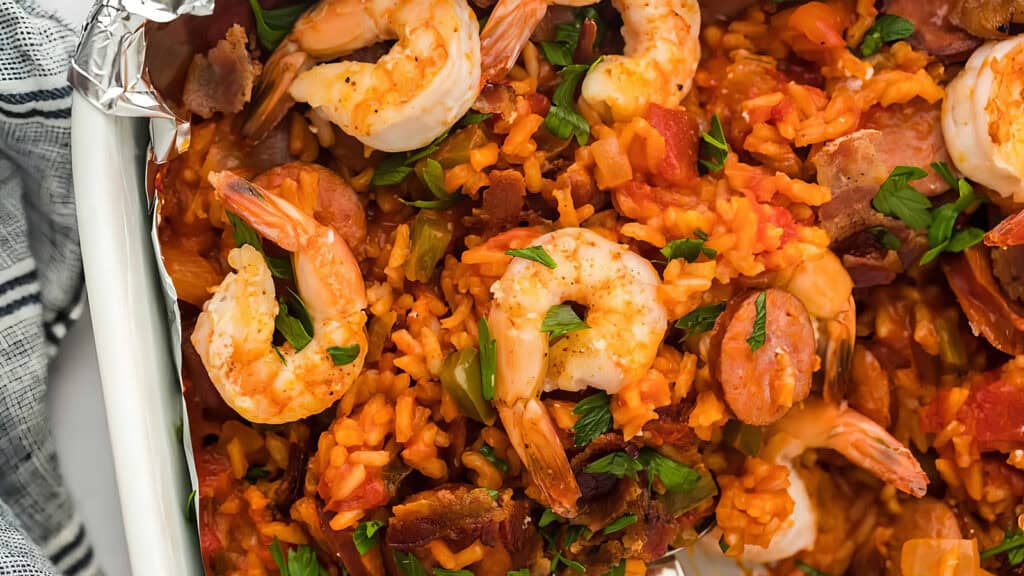 A close-up of a delicious shrimp jambalaya dish featuring plump shrimp, sausage slices, rice, diced tomatoes, and green peppers. Garnished with fresh parsley, this vibrant and colorful Southern recipe sits in a white dish next to a striped towel.