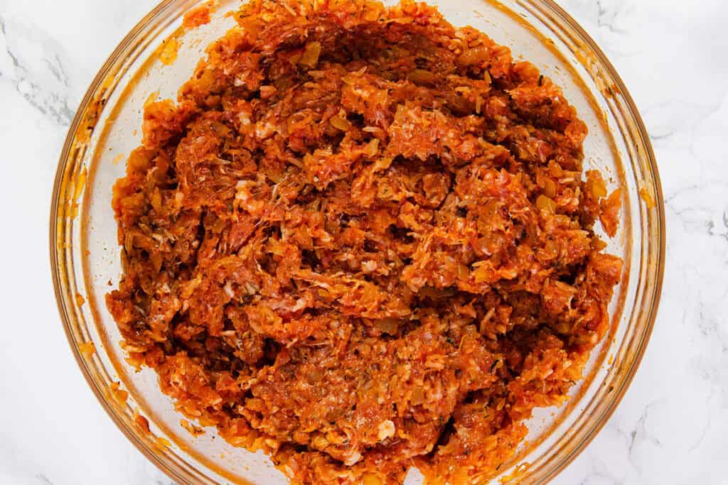 A close-up of a clear glass bowl filled with a mixture of ground meat, finely chopped onions, and spices for making Sarmale. The ingredients are well-combined, creating a reddish-orange blend with a slightly chunky texture. The bowl is placed on a white marble surface.