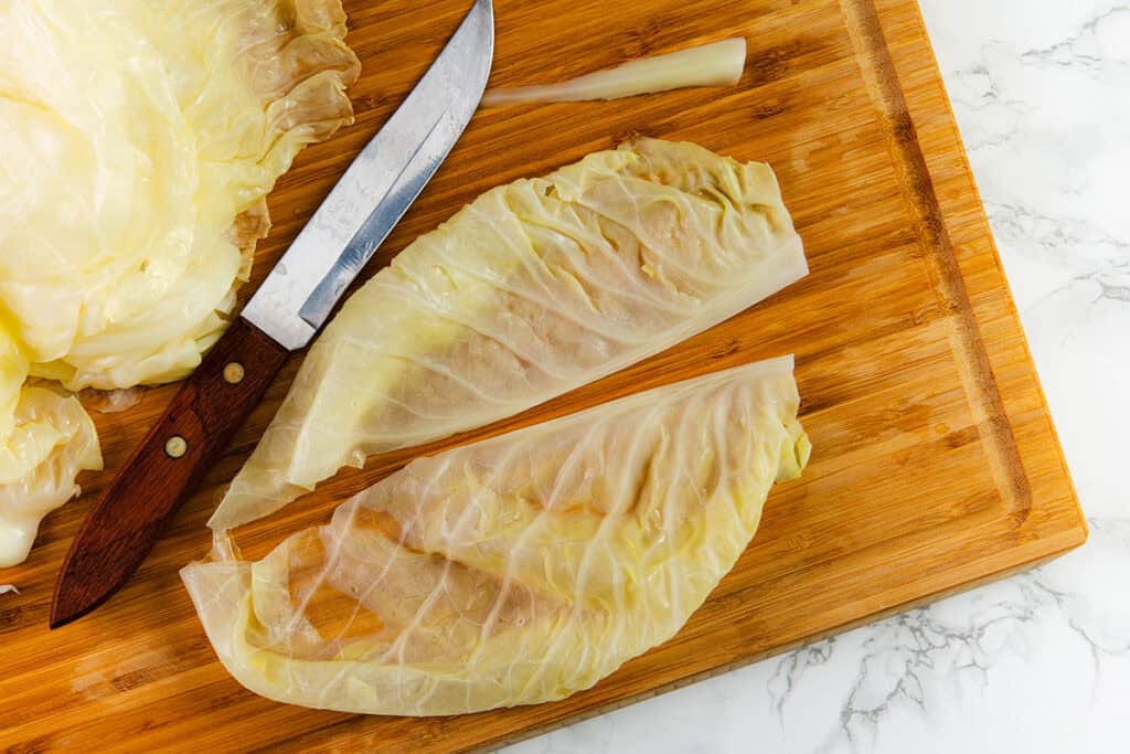 Two cabbage leaves lie on a wooden cutting board, each filled with a meat mixture reminiscent of traditional Romanian Cabbage Rolls. A knife with a wooden handle rests nearby, beside more cabbage leaves. The surface shows a light marble pattern, indicating a kitchen countertop ready for making Sarmale.