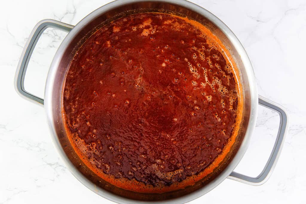 A large stainless-steel pot filled with simmering, tomato-based sauce for delicious Romanian Cabbage Rolls, also known as Sarmale. The surface of the sauce has a rich red color with visible bubbles, indicating it is being cooked. The pot has two sturdy handles on each side, set against a white marble countertop.
