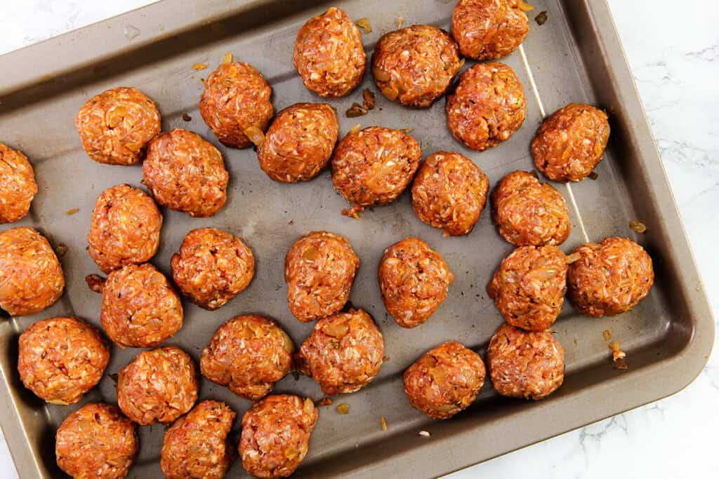 A baking tray is filled with numerous raw meatballs, evenly spaced apart. The meatballs have a rich, reddish-brown color, suggesting they are seasoned similar to the filling of traditional Romanian cabbage rolls, or sarmale. The tray sits on a white marble surface, ready to be placed in the oven.