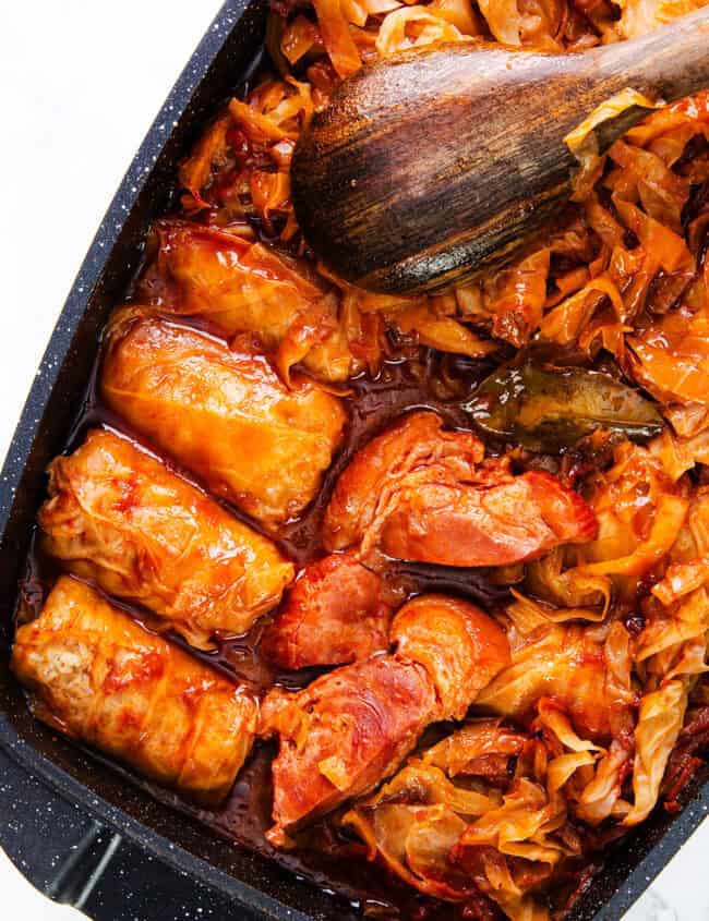 A close-up view of a pot filled with Romanian Cabbage Rolls and chunks of meat in a rich, reddish sauce. A wooden spoon is resting on top of the food, indicating a freshly prepared and hearty dish.