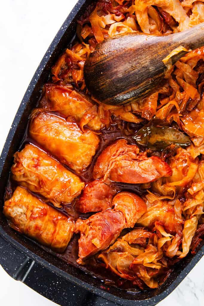 A close-up view of a pot filled with Romanian Cabbage Rolls and chunks of meat in a rich, reddish sauce. A wooden spoon is resting on top of the food, indicating a freshly prepared and hearty dish.