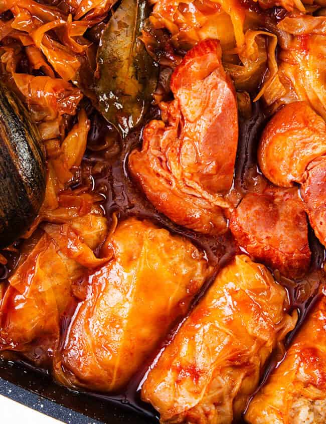 A close-up image of a cooked dish featuring stuffed cabbage rolls in a rich, reddish sauce with shredded cabbage. A wooden spoon rests on the left side of the pan, partly submerged in the sauce and cabbage mixture. Perfect for those who love slow cooker recipes, this dish promises hearty flavors and comfort.