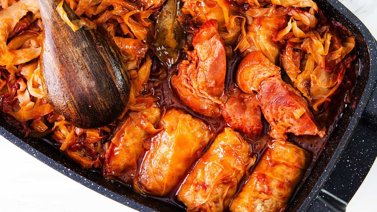 A close-up image of a cooked dish featuring stuffed cabbage rolls in a rich, reddish sauce with shredded cabbage. A wooden spoon rests on the left side of the pan, partly submerged in the sauce and cabbage mixture. Perfect for those who love slow cooker recipes, this dish promises hearty flavors and comfort.