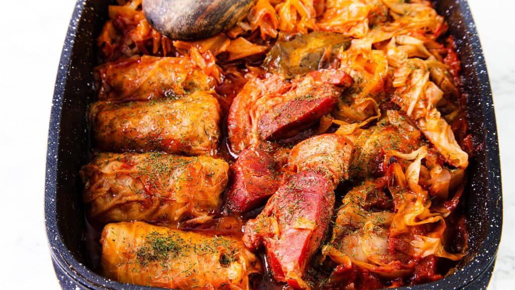 A close-up of a dish featuring Romanian Cabbage Rolls, or Sarmale, and cuts of meat, all cooked in a rich, spiced tomato sauce. The food is garnished with herbs and is served in a black, speckled baking dish. The vibrant colors of the dish make it look inviting and appetizing.