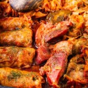 A close-up of a dish featuring Romanian Cabbage Rolls, or Sarmale, and cuts of meat, all cooked in a rich, spiced tomato sauce. The food is garnished with herbs and is served in a black, speckled baking dish. The vibrant colors of the dish make it look inviting and appetizing.