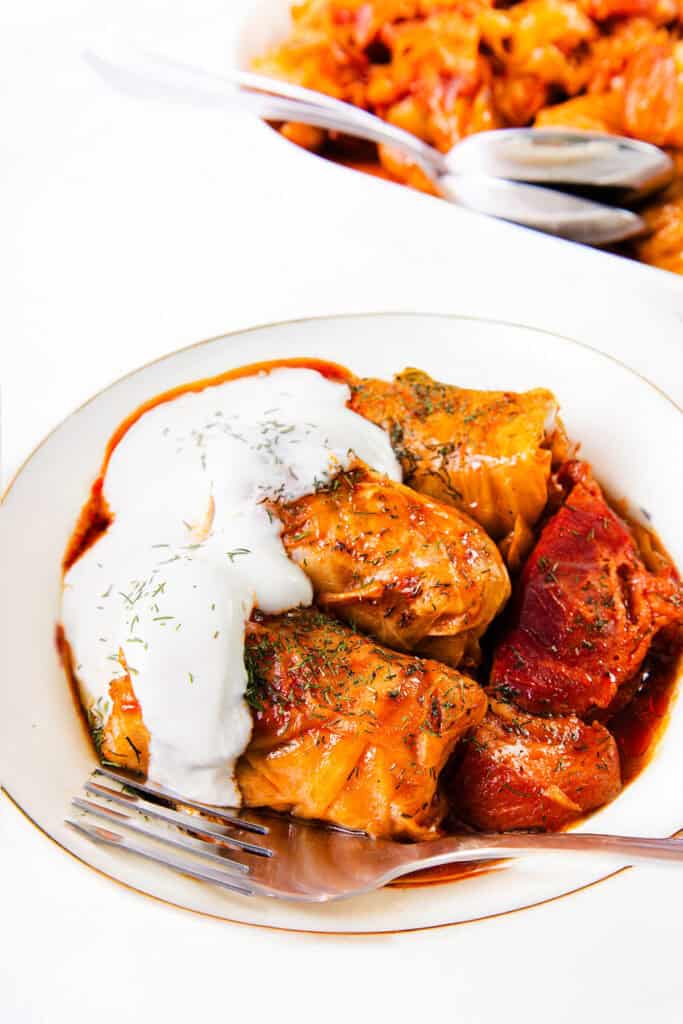 A white bowl filled with Romanian Cabbage Rolls, known as Sarmale, in tomato sauce, garnished with a generous layer of sour cream and a sprinkle of dill. A fork rests on the edge of the bowl. Another dish with more cabbage rolls and a spoon is visible in the background.