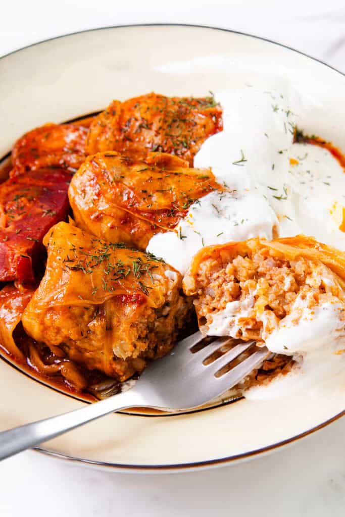 A close-up shot of a plate containing Romanian cabbage rolls, also known as sarmale, filled with rice and meat. They are topped with a tomato-based sauce and a dollop of sour cream sprinkled with dill. One roll is cut in half, revealing the filling, and a fork rests beside it.