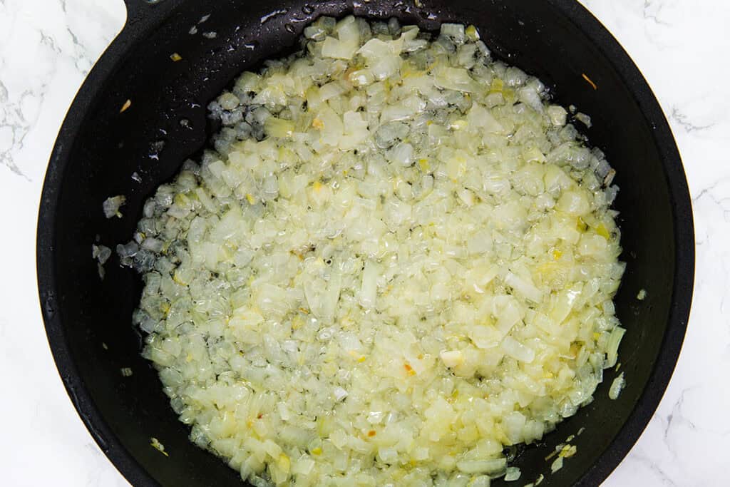 A black skillet filled with finely chopped onions being sautéed for delicious Romanian Cabbage Rolls. The translucent, light golden onions indicate they are partially cooked. The skillet is positioned on a white marble countertop, preparing the flavorful base for sarmale.