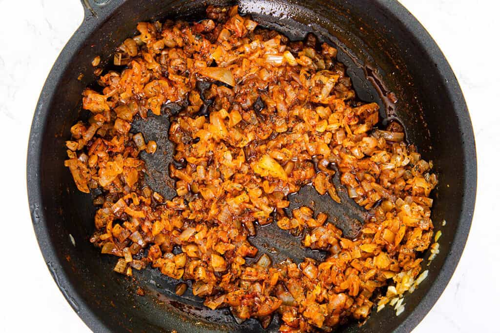 A black skillet containing a mixture of sautéed diced onions and garlic with a rich orange-red hue, indicating the presence of spices such as paprika or chili powder, and a slightly caramelized surface on some of the pieces. The pan, perfect for starting off Romanian sarmale (cabbage rolls), is placed on a marble countertop.