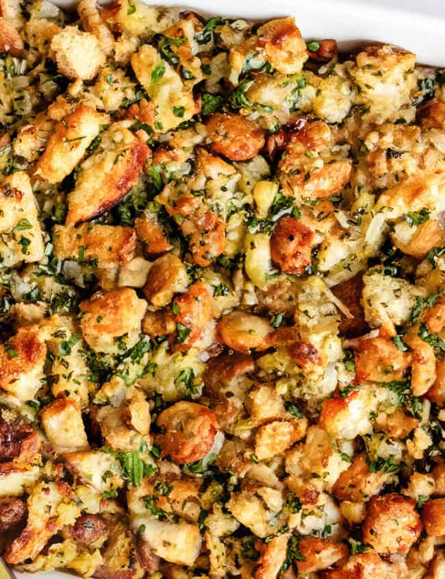 A close-up of a baking dish filled with golden-brown stuffing, a quintessential Thanksgiving side. The stuffing consists of chunks of bread, chopped vegetables, and herbs, all mixed together and baked to a crispy texture on top. A spoon is partially submerged in the dish, ready to serve.