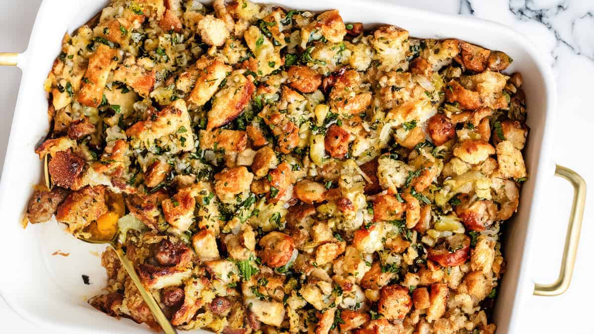 A close-up of a baking dish filled with golden-brown stuffing, a quintessential Thanksgiving side. The stuffing consists of chunks of bread, chopped vegetables, and herbs, all mixed together and baked to a crispy texture on top. A spoon is partially submerged in the dish, ready to serve.