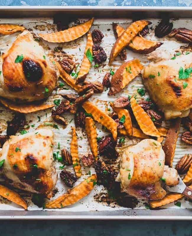 A baking tray filled with roasted chicken thighs, sweet potato wedges, pecans, and garnished with fresh herbs. The food is golden-brown and appears savory, perfect for an easy weeknight dinner, with a blue textured backdrop. A wooden spoon rests on the tray.