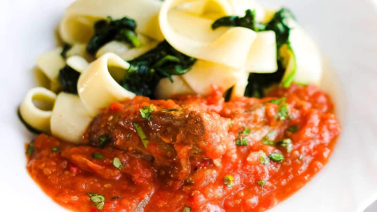 A plate of pasta with wide noodles, wilted greens, and a hearty tomato sauce reminiscent of a classic stew. The chunky sauce boasts visible pieces of herbs and generous portions of meat. The greens are interspersed among the noodles, giving the dish a freshly made appearance perfect for any cooking enthusiast.