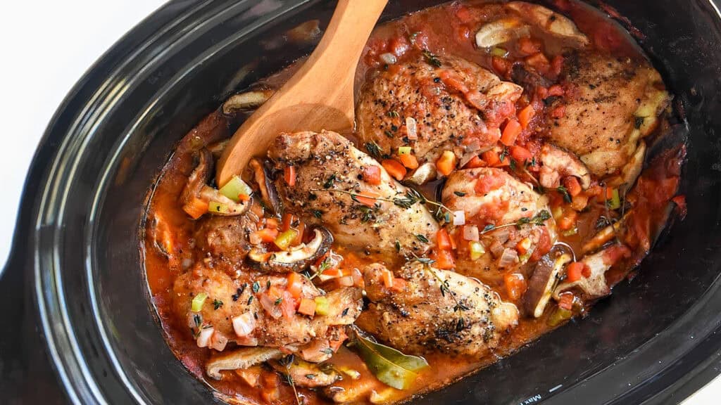 A close-up of a slow cooker filled with a rich, hearty dish. The meal consists of seasoned chicken thighs surrounded by chopped vegetables, including bell peppers and onions, all nestled in a savory, tomato-based broth. A wooden spoon is stirring the contents—a perfect inspiration for slow cooker recipes.