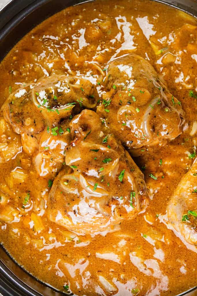 A close-up of a rich, creamy curry dish featuring multiple pieces of tender smothered chicken. The curry sauce is thick and glossy, garnished with chopped herbs, and contains visible slices of onions and spices, suggesting a flavorful and hearty meal.