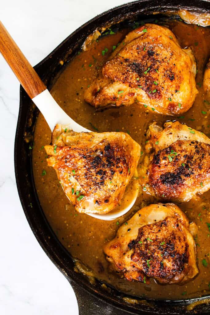 A skillet filled with golden-brown, crispy chicken thighs partially submerged in a rich, brown gravy. A wooden-handled spatula is lifting one of the smothered chicken thighs out of the skillet. The dish is garnished with finely chopped herbs.