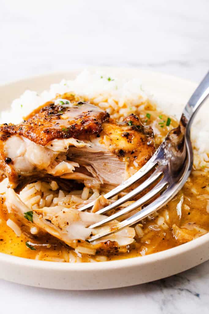 A fork cuts into a piece of grilled chicken thigh served over a bed of white rice, smothered in a yellow sauce that has herbs and oil droplets. The chicken is juicy and browned, and the rice absorbs some of the sauce, adding extra flavor.