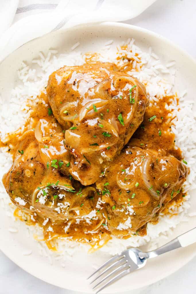 A plate with three pieces of smothered chicken covered in a savory brown gravy, garnished with chopped herbs, is served on a bed of white rice. A fork rests on the edge of the plate.