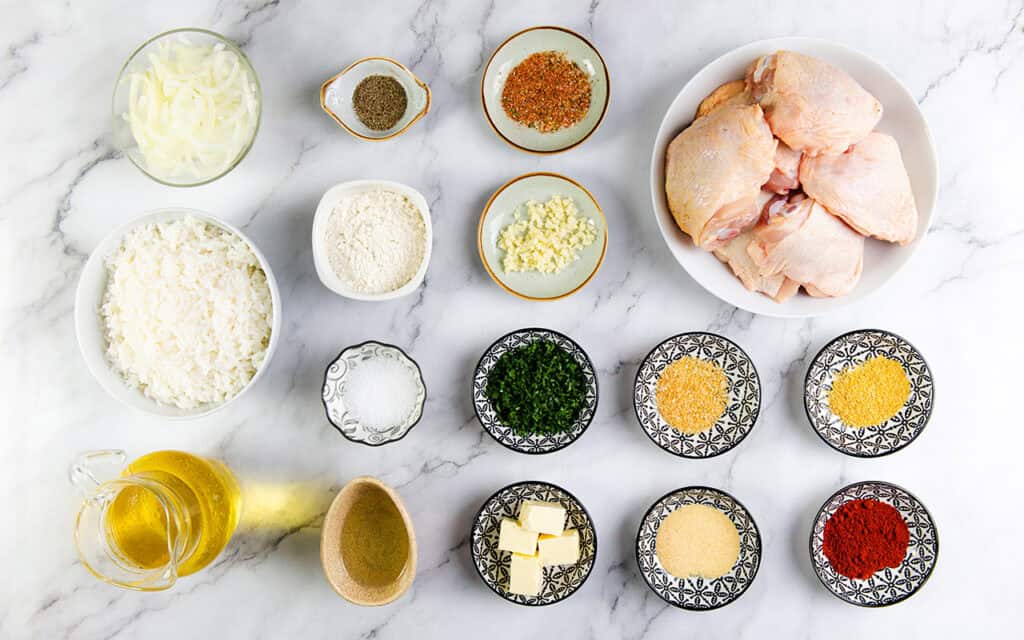 Top-down view of various cooking ingredients arranged on a marble surface, including raw chicken pieces for a smothered chicken thighs recipe, sliced onions, minced garlic, diced herbs, spices in small bowls, flour, rice, butter, oil, and a liquid gravy seasoning in a small pitcher.