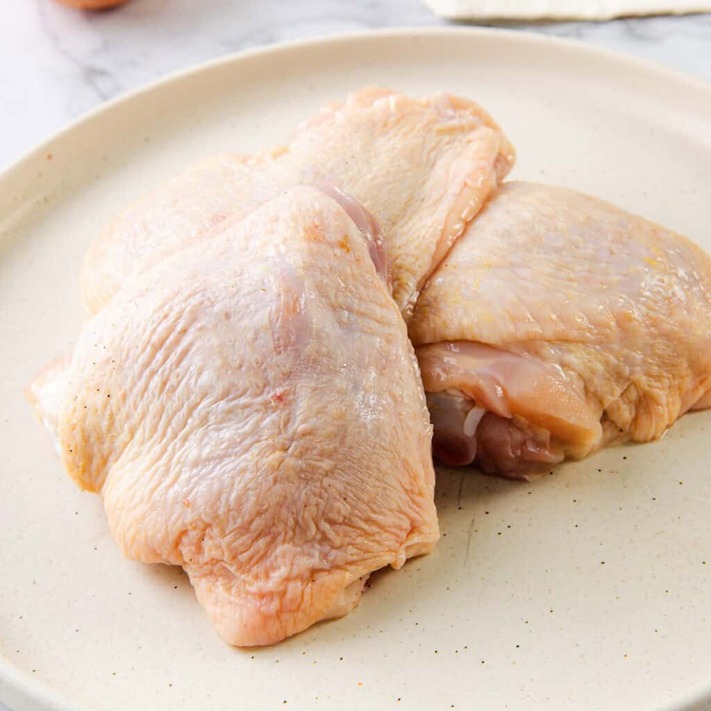 A plate with three raw chicken thighs with skin on. The chicken is pale pink, and the plate is off-white, resting on a white marble surface. The chicken appears fresh and uncooked, ready for preparation into a delicious smothered chicken with gravy.