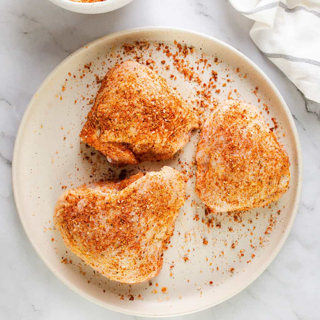 A plate with three smothered chicken thighs covered with a blend of spices. The plate is set on a marble surface, with a striped dish towel partially visible on the right and a small bowl in the upper left corner.