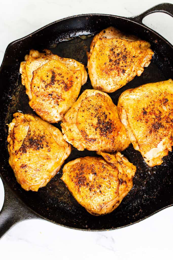 A cast-iron skillet filled with six golden brown, crispy smothered chicken thighs, seasoned with visible black pepper and other spices. The skillet rests on a white surface, showcasing the contrast between the dark pan and the vibrant, well-cooked chicken pieces.