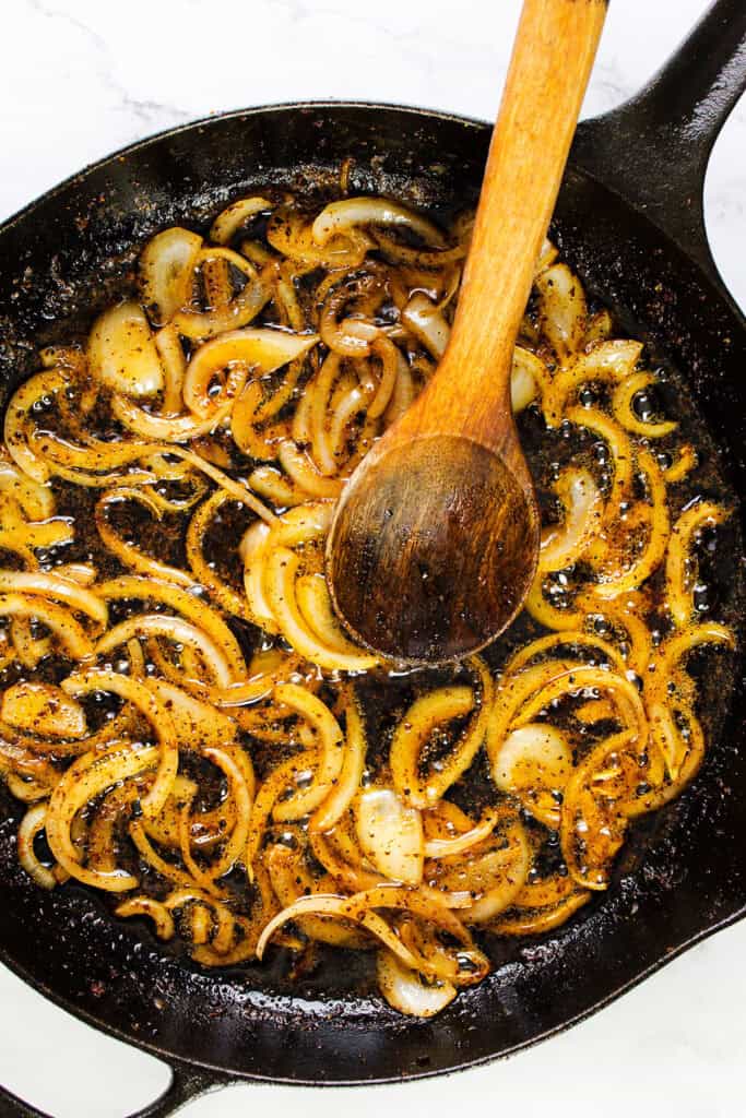 A cast iron skillet containing caramelized onions being stirred with a wooden spoon. The onions are golden brown, seasoned with pepper, and nestled in a mix of oil and seasoning, giving off a savory appearance, perfect for smothered chicken.