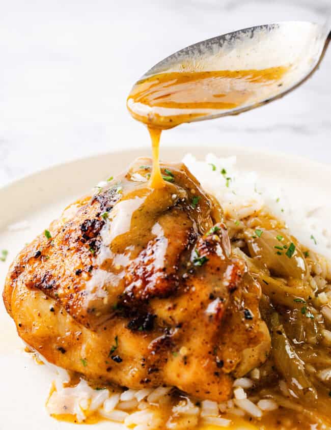 A savory serving of smothered chicken thighs covered in a rich brown gravy sauce sits on a plate with white rice. A spoon is held above, drizzling more gravy onto the tender chicken. Garnish of finely chopped herbs adds a touch of green to the dish.