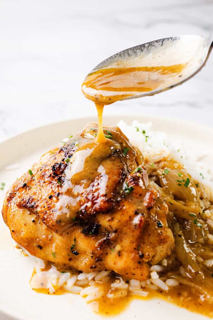 A savory serving of smothered chicken thighs covered in a rich brown gravy sauce sits on a plate with white rice. A spoon is held above, drizzling more gravy onto the tender chicken. Garnish of finely chopped herbs adds a touch of green to the dish.