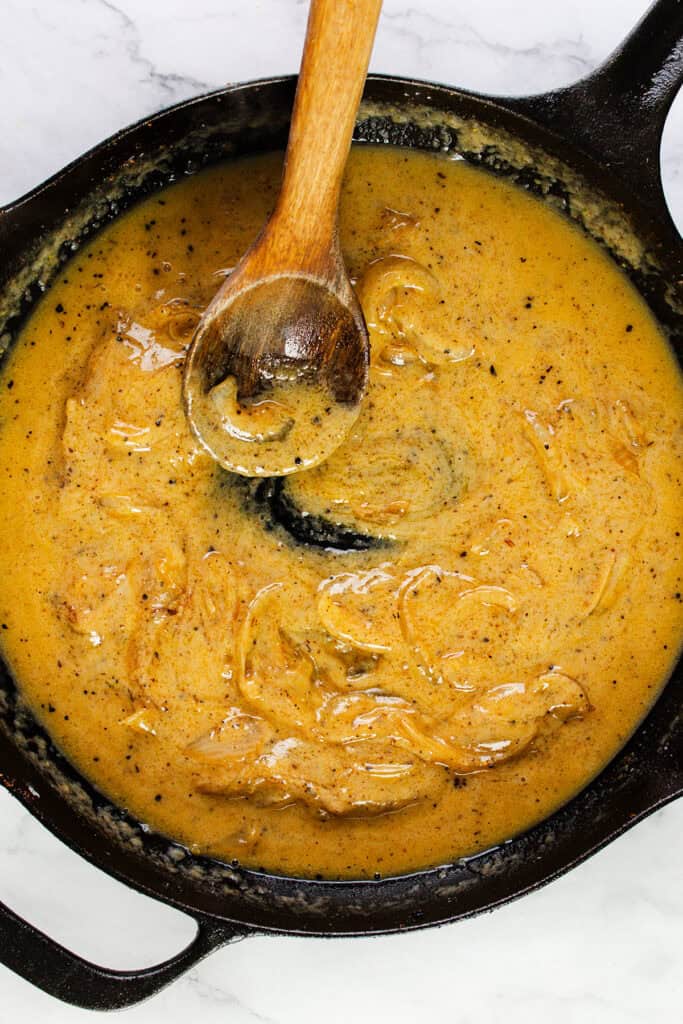 A cast iron skillet filled with creamy, golden-yellow onion gravy, perfect for smothered chicken. A wooden spoon rests in the center, partially submerged in the thick sauce. The gravy has visible onion pieces and is speckled with black pepper, all set on a marble countertop.