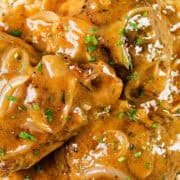 A close-up of a plate of smothered chicken covered in a creamy brown sauce with sliced onions, garnished with chopped herbs, and served over a bed of white rice. A fork is partially visible on the right side of the plate.