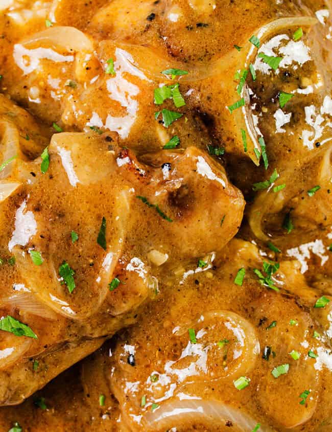 A close-up of a plate of smothered chicken covered in a creamy brown sauce with sliced onions, garnished with chopped herbs, and served over a bed of white rice. A fork is partially visible on the right side of the plate.