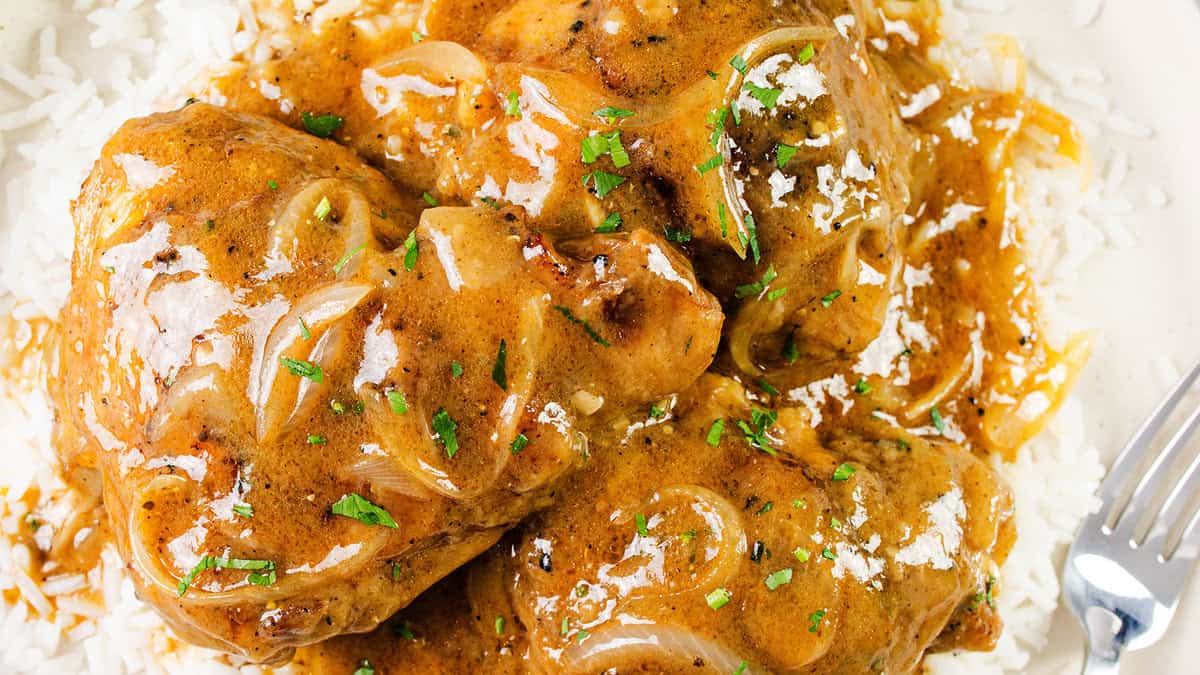 A close-up of a plate of smothered chicken covered in a creamy brown sauce with sliced onions, garnished with chopped herbs, and served over a bed of white rice. A fork is partially visible on the right side of the plate.