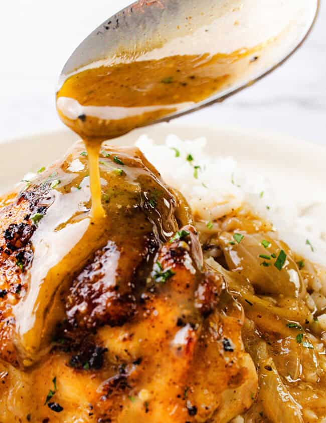 A close-up of a plate with grilled chicken, caramelized onions, and white rice, garnished with chopped herbs. A spoon is drizzling a golden-brown sauce over the chicken, creating a savory and appetizing presentation. This easy chicken dinner idea is sure to impress every time on your marble-like surface.