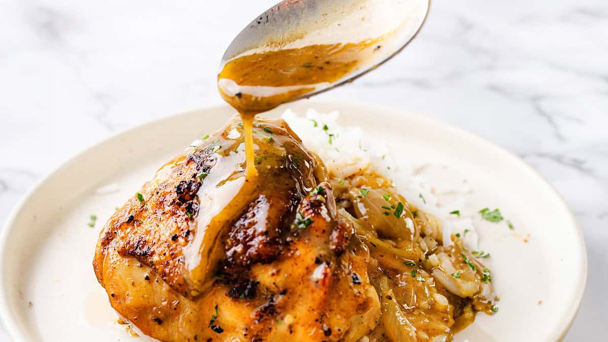 A close-up of a plate with grilled chicken, caramelized onions, and white rice, garnished with chopped herbs. A spoon is drizzling a golden-brown sauce over the chicken, creating a savory and appetizing presentation. This easy chicken dinner idea is sure to impress every time on your marble-like surface.