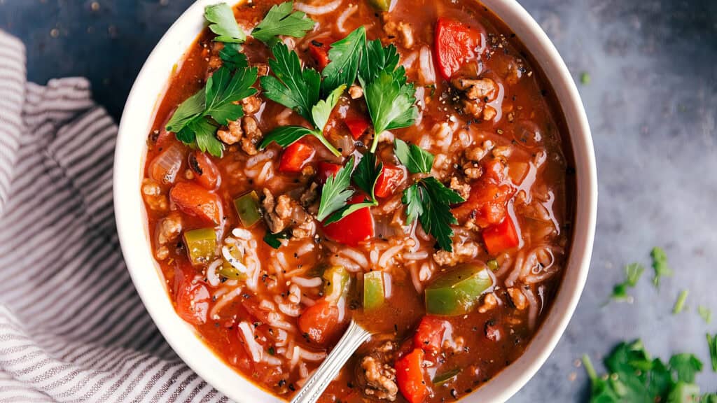 A bowl of hearty soup filled with ground beef, rice, tomatoes, green and red bell peppers, garnished with fresh parsley. A spoon rests in the soup, and a striped cloth napkin is visible beside the bowl, all set against a dark textured background — perfect for ground beef recipes.