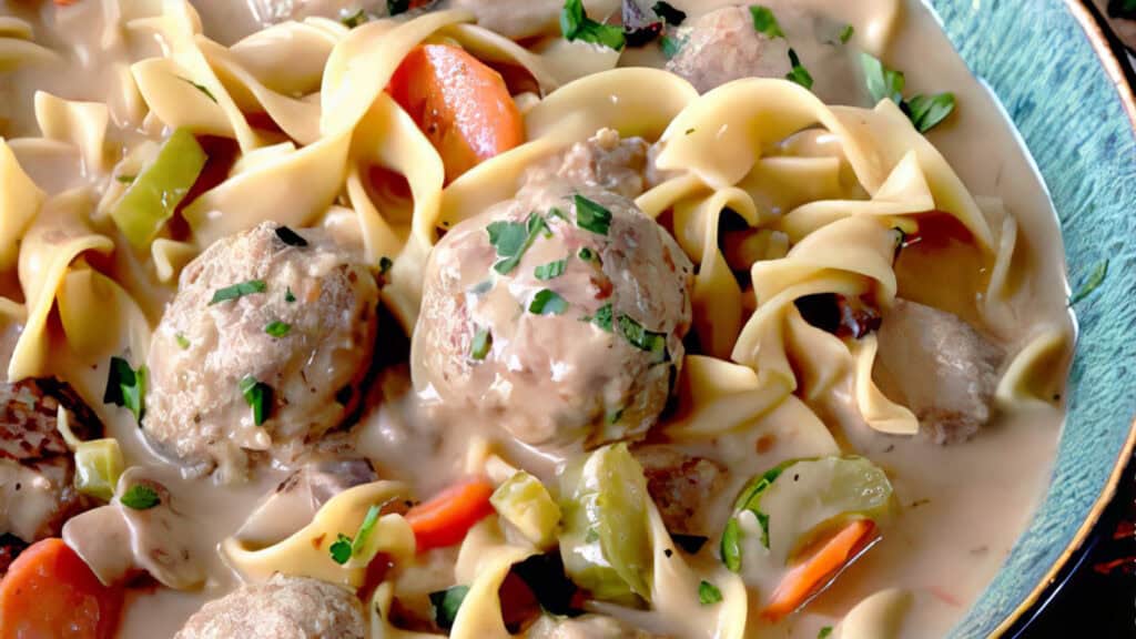 A close-up of a bowl filled with creamy chicken and vegetable noodle soup. The soup contains egg noodles, chicken chunks, meatballs made from ground beef recipes, carrots, celery, and is garnished with chopped fresh herbs. The rich broth looks smooth and hearty.