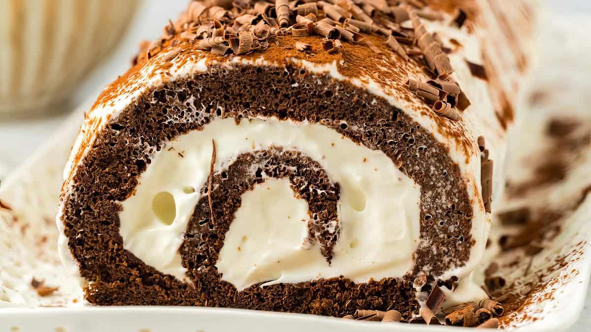 A close-up of a chocolate Swiss roll cake, showcasing a spiral of rich chocolate sponge wrapped around a creamy white filling. The cake is topped with a dusting of cocoa powder and chocolate shavings, highlighting its moist and indulgent texture.